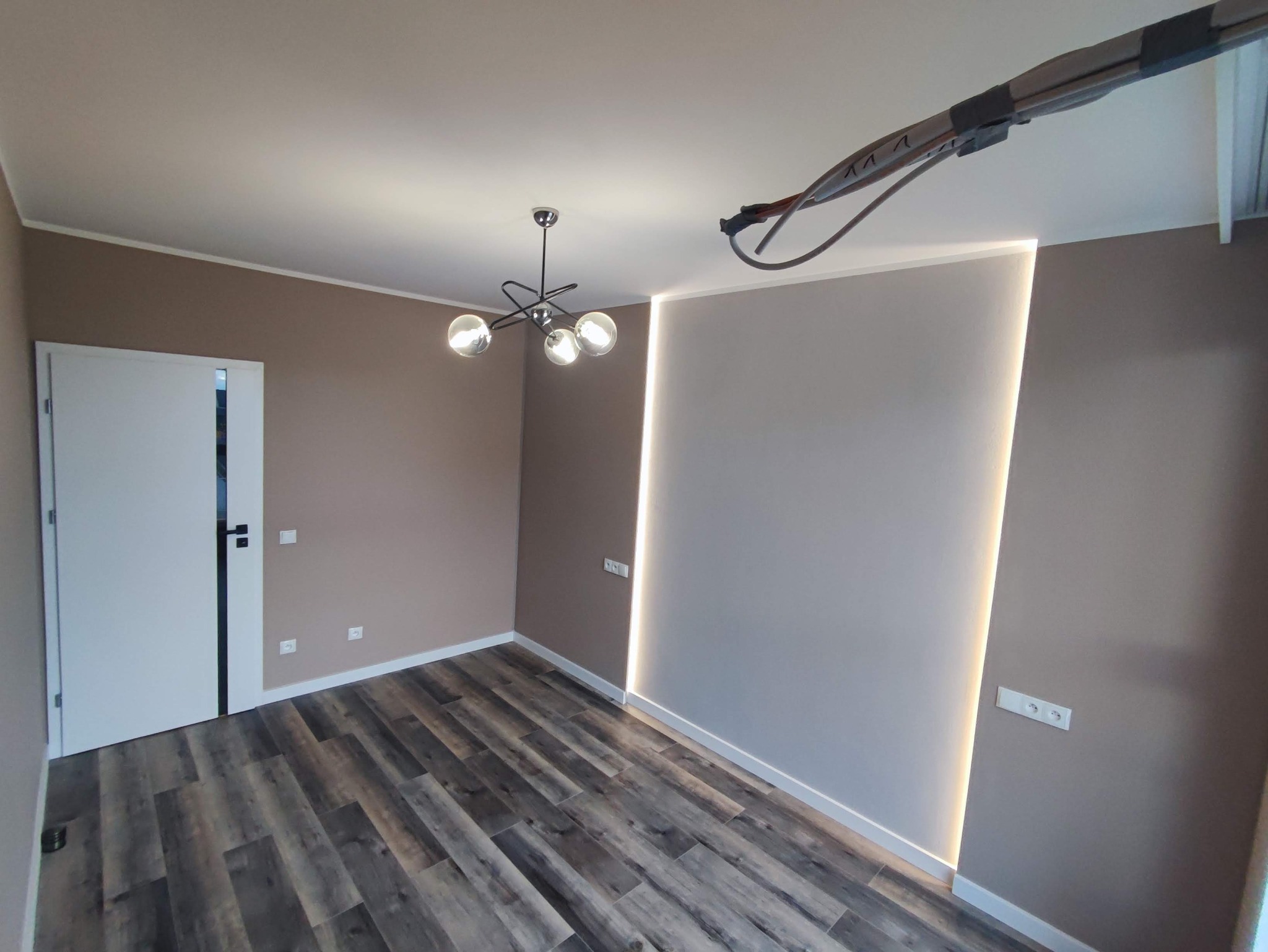 Wnętrze pokoju po odświeżeniu: brązowe ściany, ciemne panele podłogowe, nowoczesna lampa sufitowa z kulistymi kloszami, pionowe oświetlenie LED w ścianie, białe listwy przypodłogowe i białe drzwi...