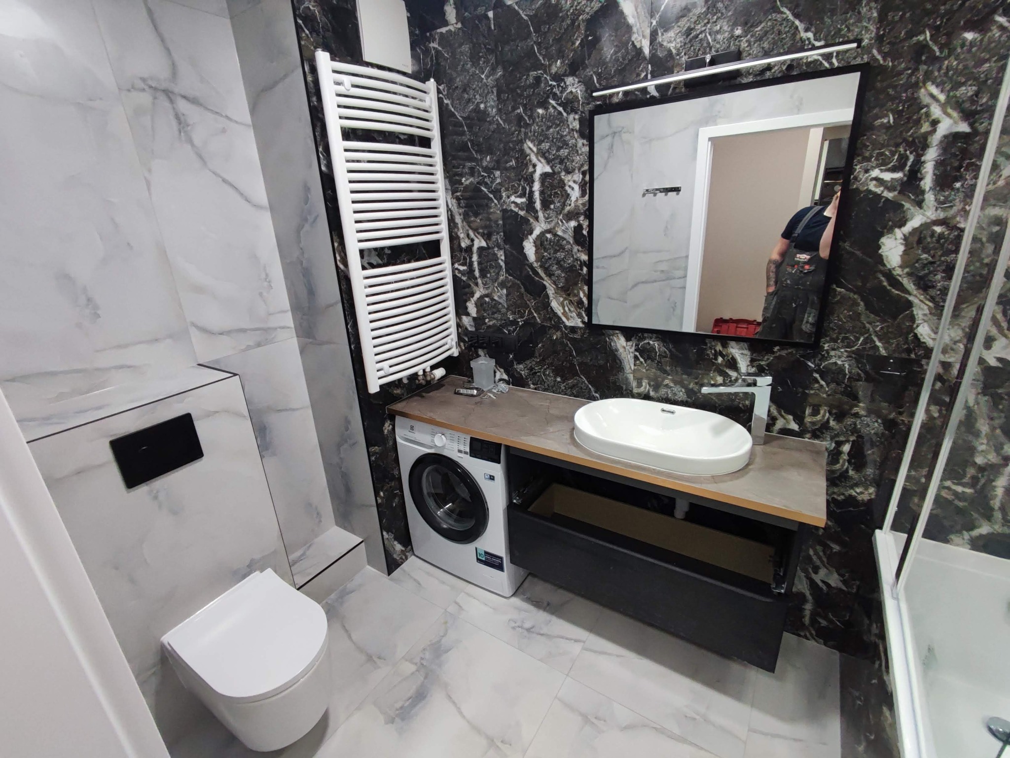 Nowoczesna łazienka z marmurowymi płytkami w odcieniach bieli i czerni, wisząca toaleta, pralka pod blatem z umywalką nablatową, grzejnik drabinkowy.