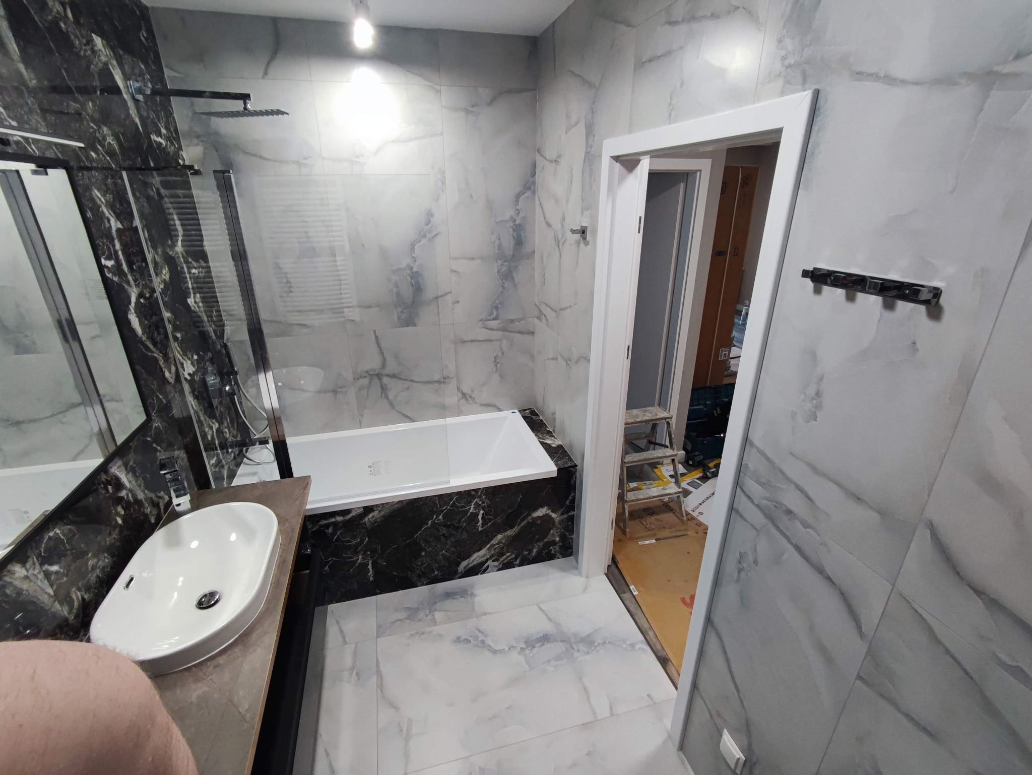 Wykończona łazienka z wanną obudowaną czarnym marmurem, białą umywalką nablatową i płytkami imitującymi marmur na ścianach i podłodze, widoczna drabina malarska w przejściu do innego pomieszczenia.