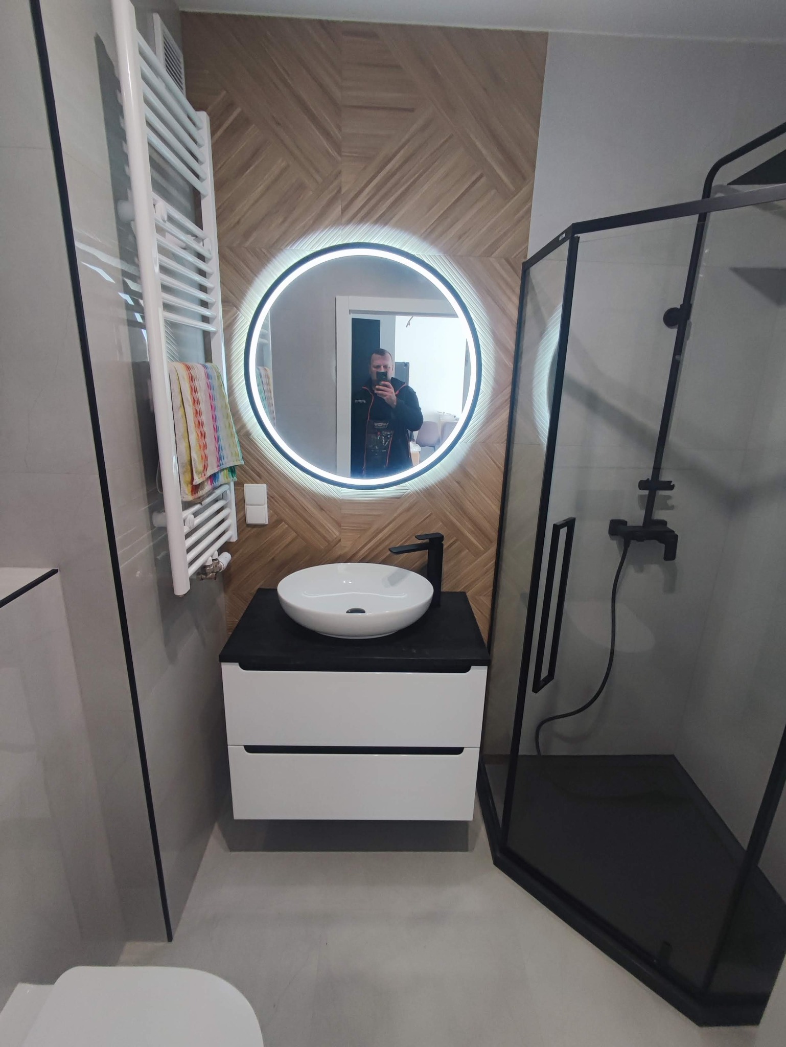 Nowoczesna łazienka z umywalką nablatową, podświetlanym lustrem, grzejnikiem drabinkowym i kabiną prysznicową z czarnymi elementami, wykończona płytkami imitującymi drewno.