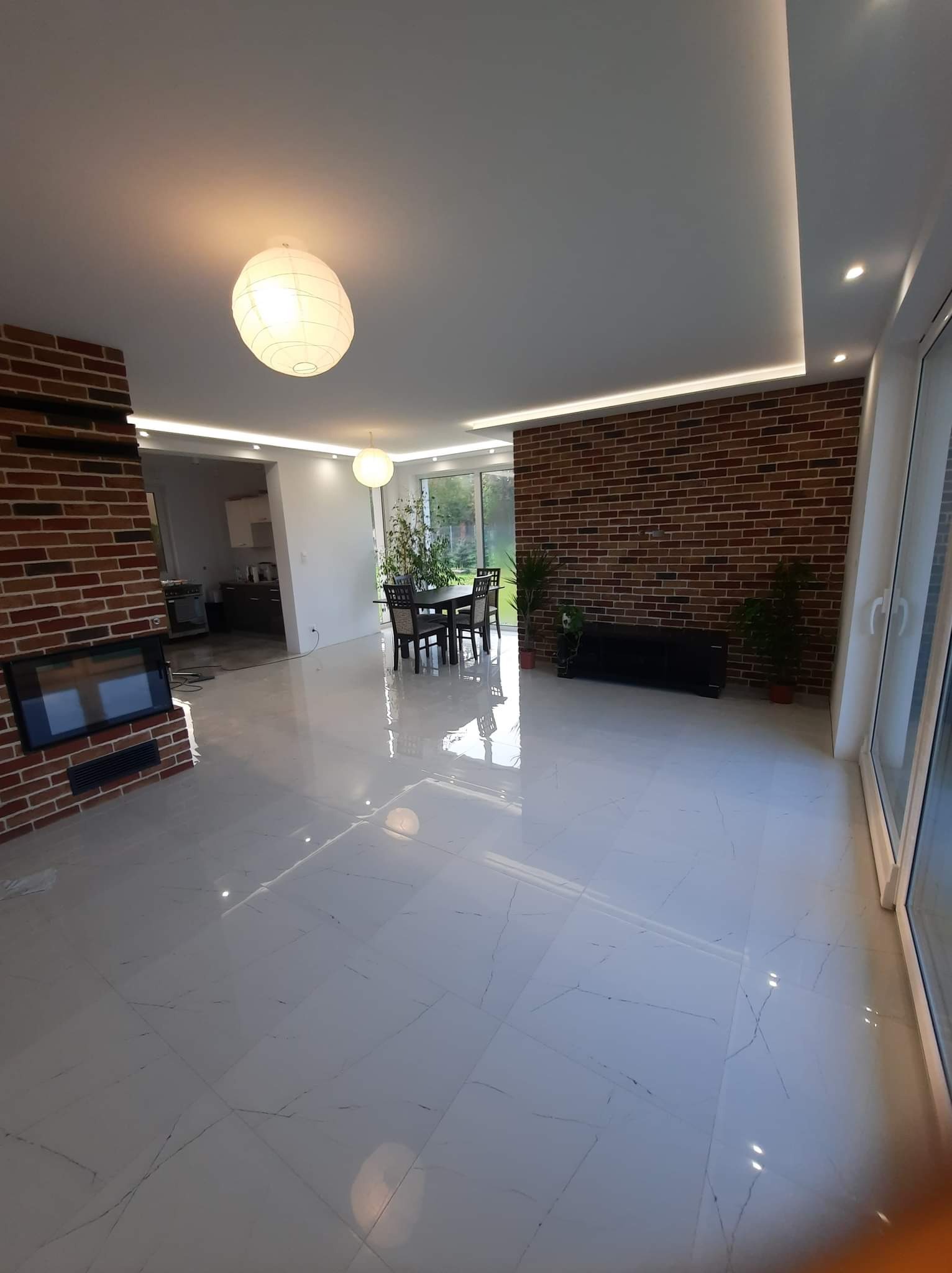 Przestronne wnętrze z błyszczącą, białą podłogą z płytek, ceglaną ścianą i kominkiem, oświetlone kulistymi lampami i listwami LED pod sufitem.