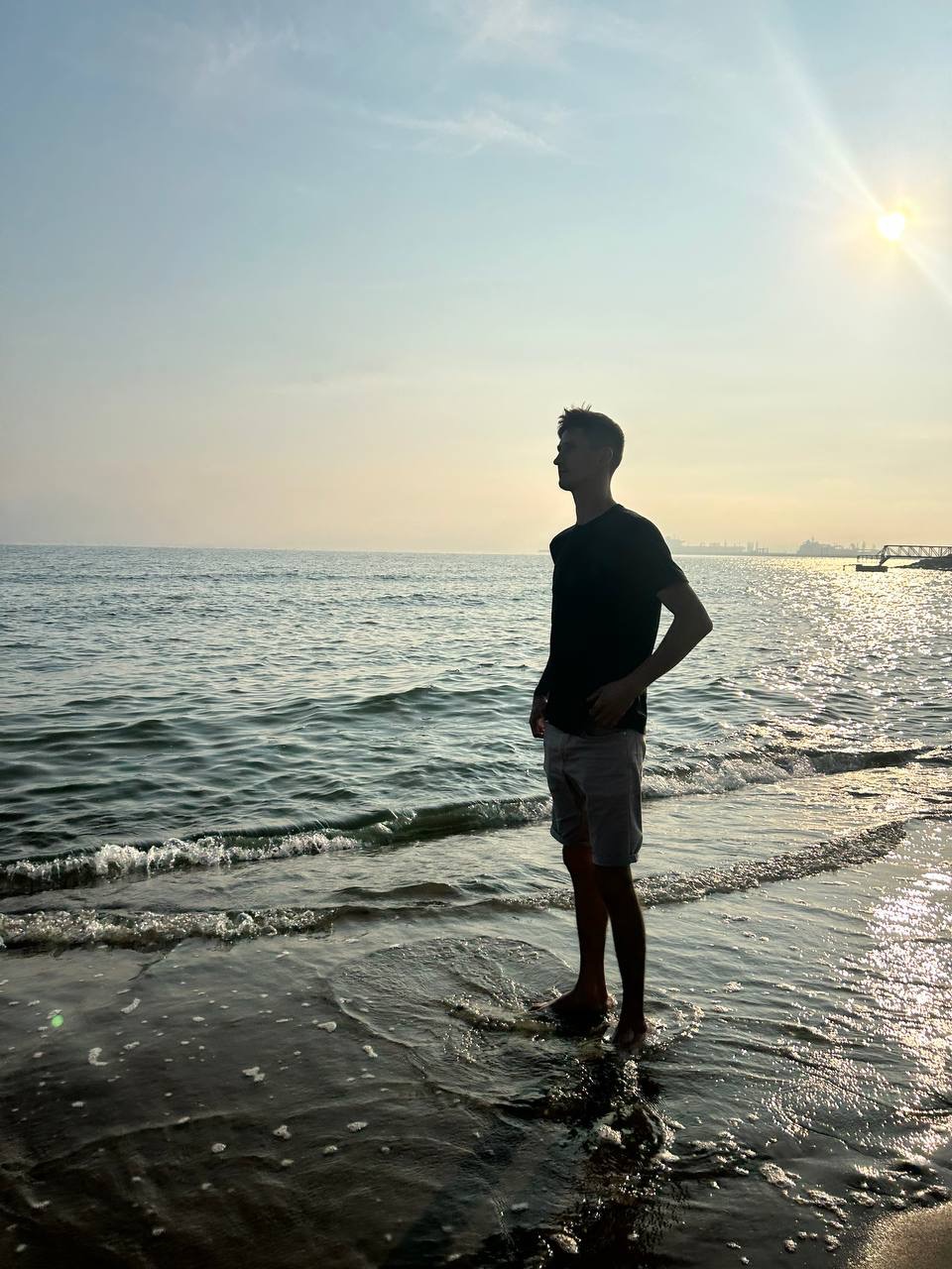 Sylwetka mężczyzny stojącego w płytkiej wodzie na brzegu morza, z widokiem na odległy horyzont i jasne słońce.