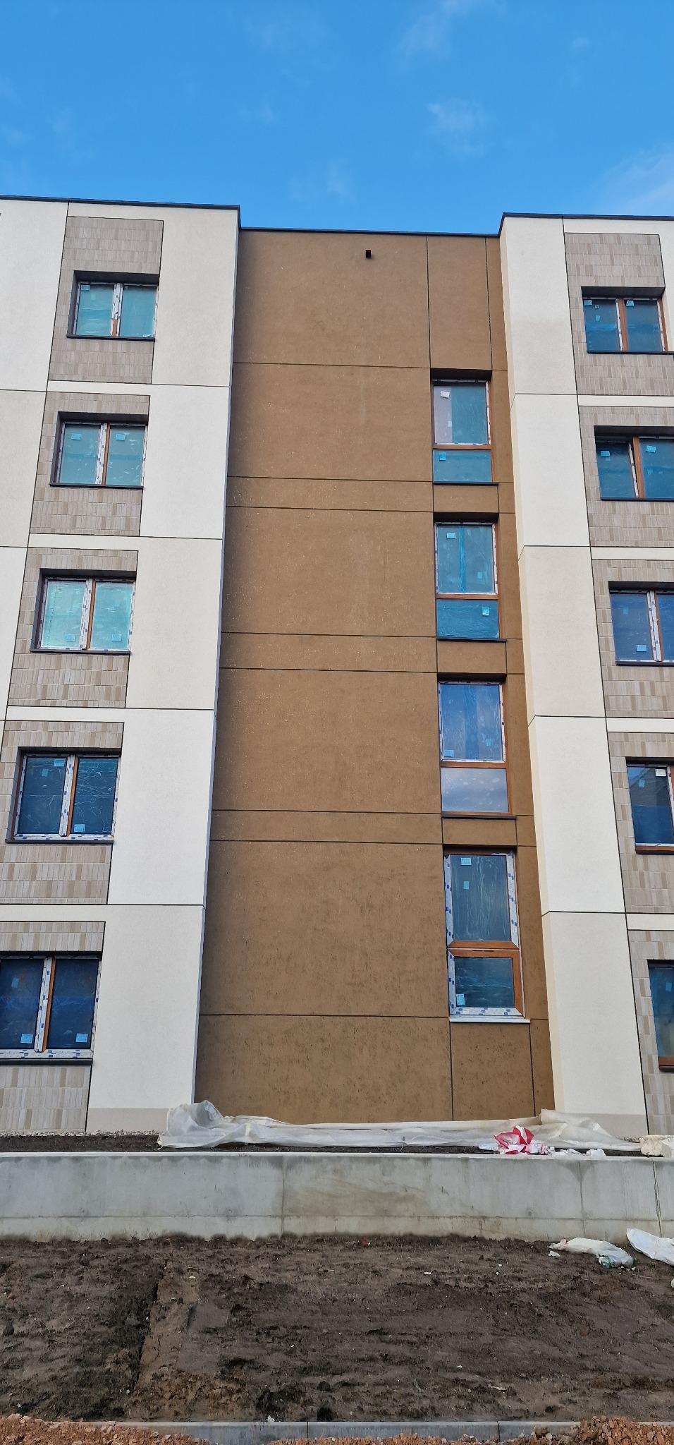 Wielopiętrowy budynek z fragmentami elewacji w kolorze beżowym i brązowym, okna zabezpieczone folią, widoczne prace wykończeniowe na dolnej kondygnacji.