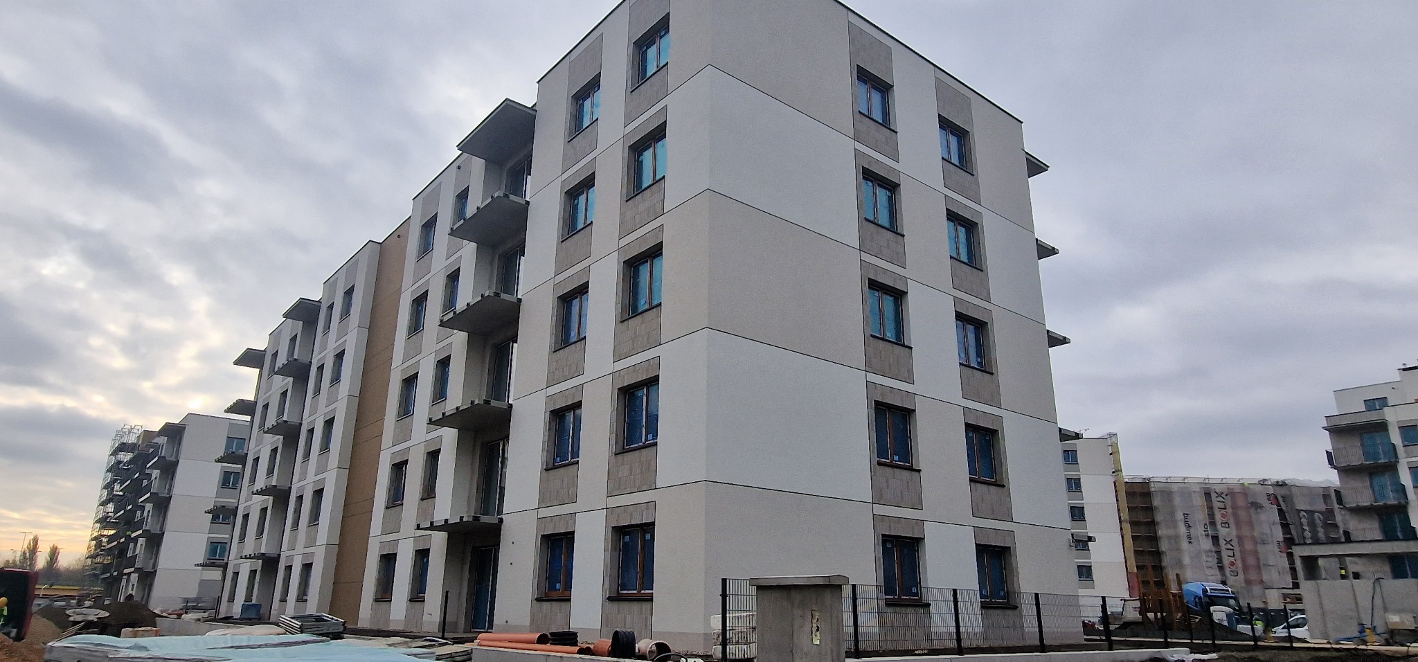 Wielopiętrowy budynek mieszkalny z jasną elewacją, widoczne balkony i okna zabezpieczone folią, na pierwszym planie materiały budowlane i ogrodzenie, w tle inne budynki w trakcie budowy.