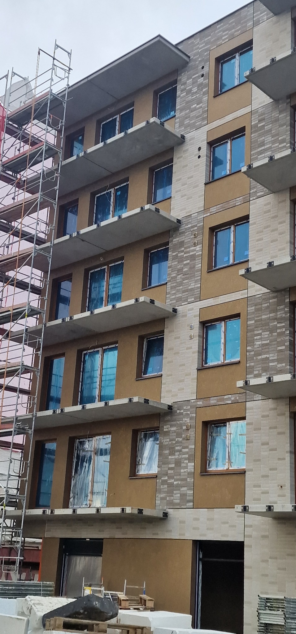 Wielopiętrowy budynek w trakcie prac wykończeniowych elewacji, z widocznymi rusztowaniami i zabezpieczonymi oknami folią, na elewacji fragmenty tynku i okładziny.
