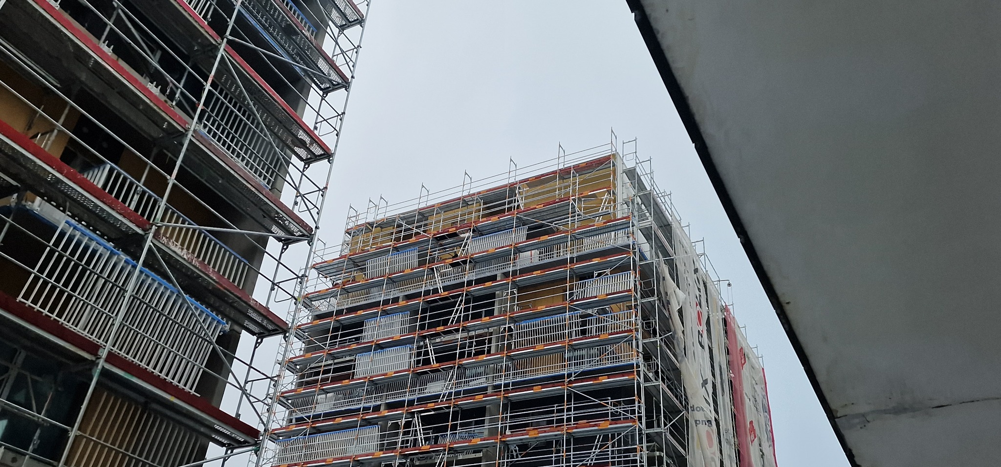 Budynek wielorodzinny w trakcie prac elewacyjnych, z widocznymi rusztowaniami i częściowo ocieploną ścianą, zabezpieczoną siatką ochronną.