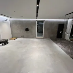 Wnętrze w trakcie remontu, widoczna część podłogi z ułożonymi panelami w jodełkę, przygotowane podłoże pod dalsze prace, paczki z materiałami budowlanymi przy ścianie, nowoczesne oświetlenie sufitowe.