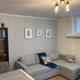 Fix PDP - Szary narożnik z turkusowym pledem i poduszkami w kwiaty na tle szaro-zielonej ściany z trzema obrazkami botanicznymi, oświetlony lampą z żarówkami Edisona.