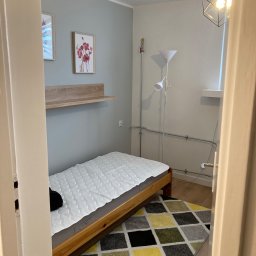 Fix PDP - Pokój po remoncie: Ściany pomalowane na szaro, drewniana półka pod obrazami, drewniane łóżko z białym materacem, wzorzysty dywan, lampa wisząca z geometrycznym kloszem, lampa stojąca z białym...