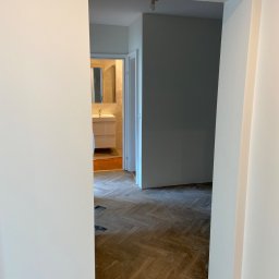 Fix PDP - Widok przez odnowione drzwi na fragment pokoju z parkietem w jodełkę, świeżo pomalowane ściany na biało i łazienkę w tle.