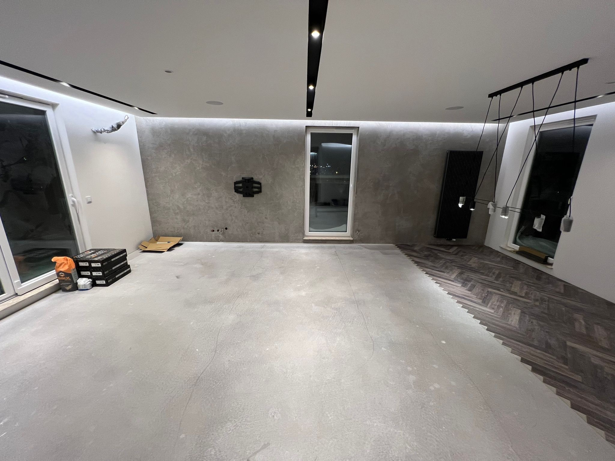 Wnętrze w trakcie remontu, widoczna część podłogi z ułożonymi panelami w jodełkę, przygotowane podłoże pod dalsze prace, paczki z materiałami budowlanymi przy ścianie, nowoczesne oświetlenie sufitowe.