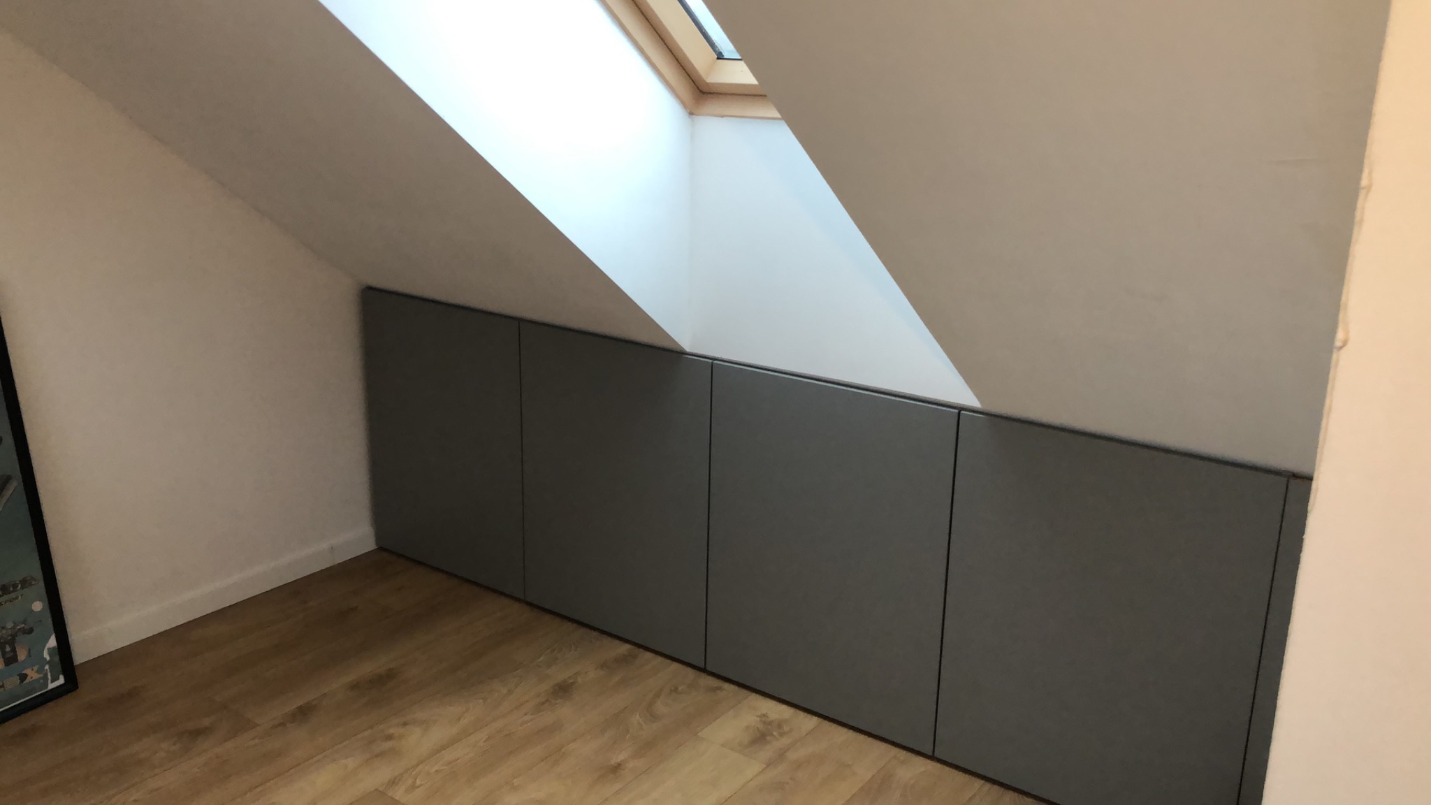 Szare zabudowane szafy pod skosem dachu, drewniana podłoga, okno dachowe wpadające światło.
