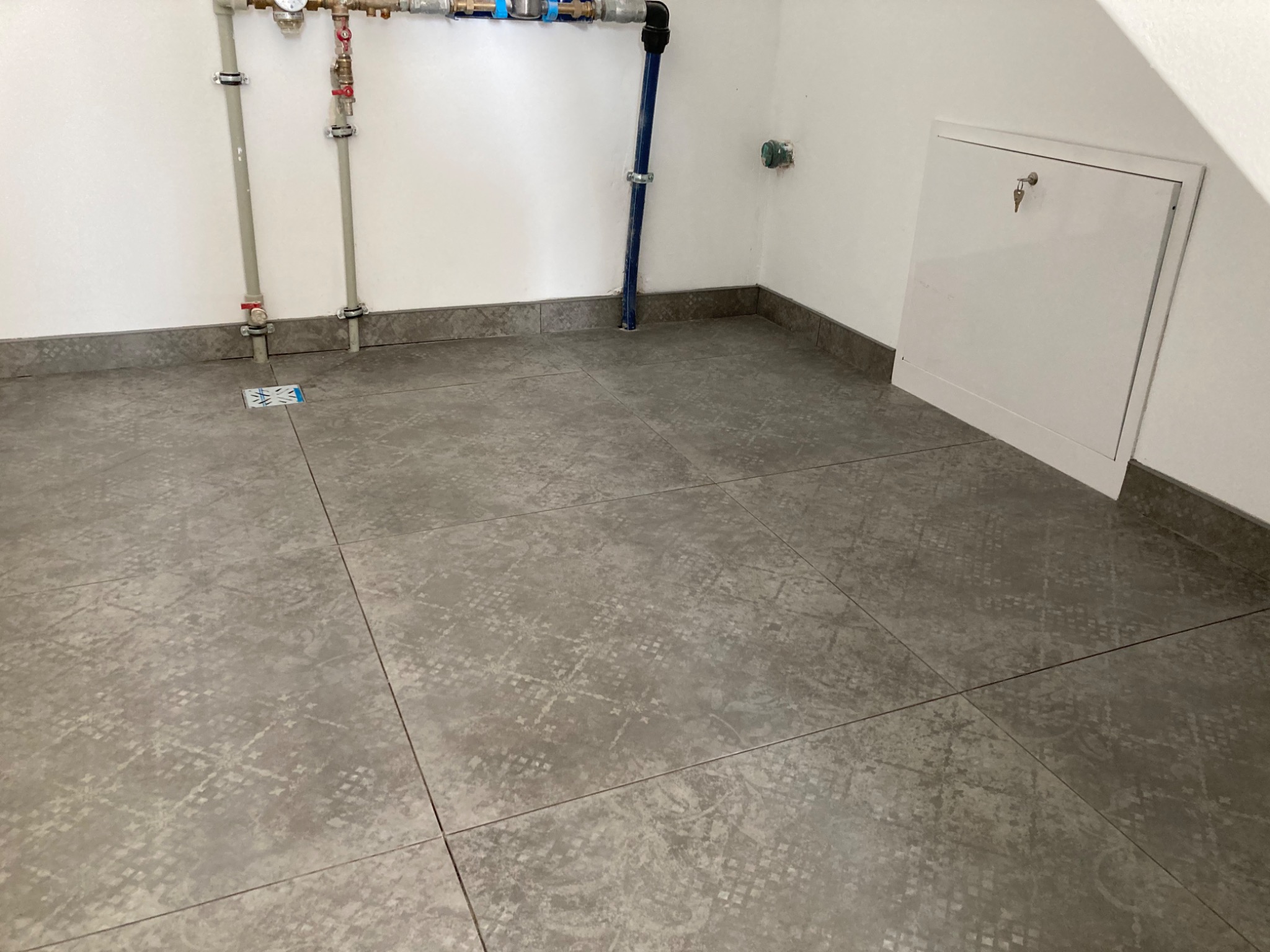 Szare płytki podłogowe z delikatnym wzorem, ułożone w pomieszczeniu z białymi ścianami, widoczne rury instalacyjne i klapka rewizyjna z kluczem.