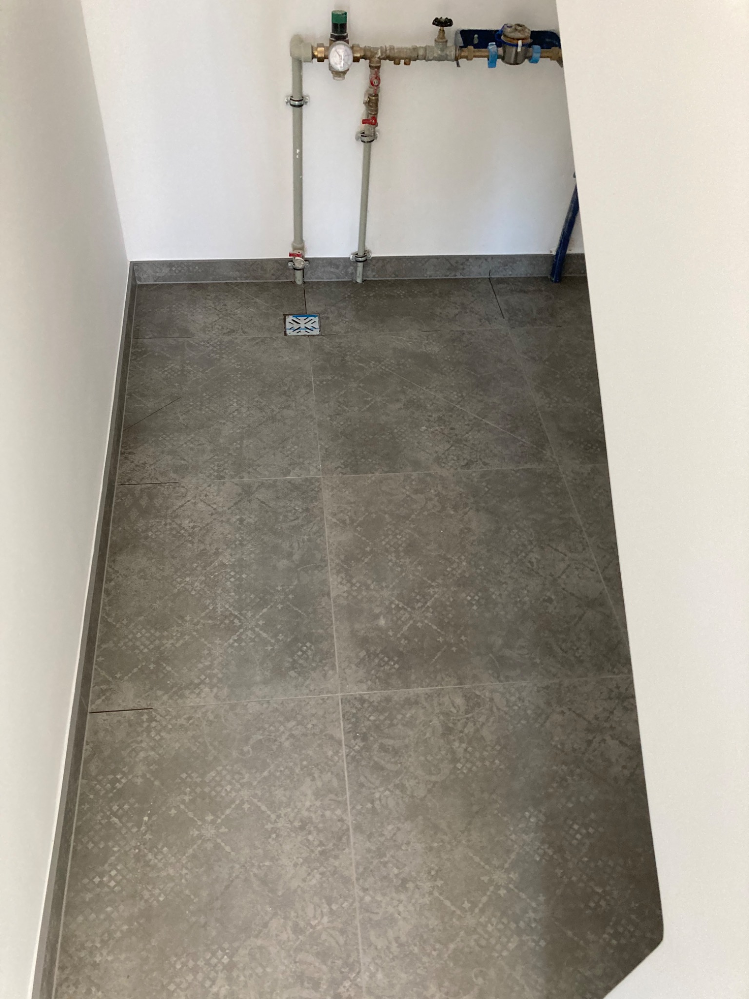 Wykończona łazienka z szarymi płytkami podłogowymi o subtelnym wzorze, widoczne rury i zawory wodne przy ścianie oraz odpływ podłogowy.