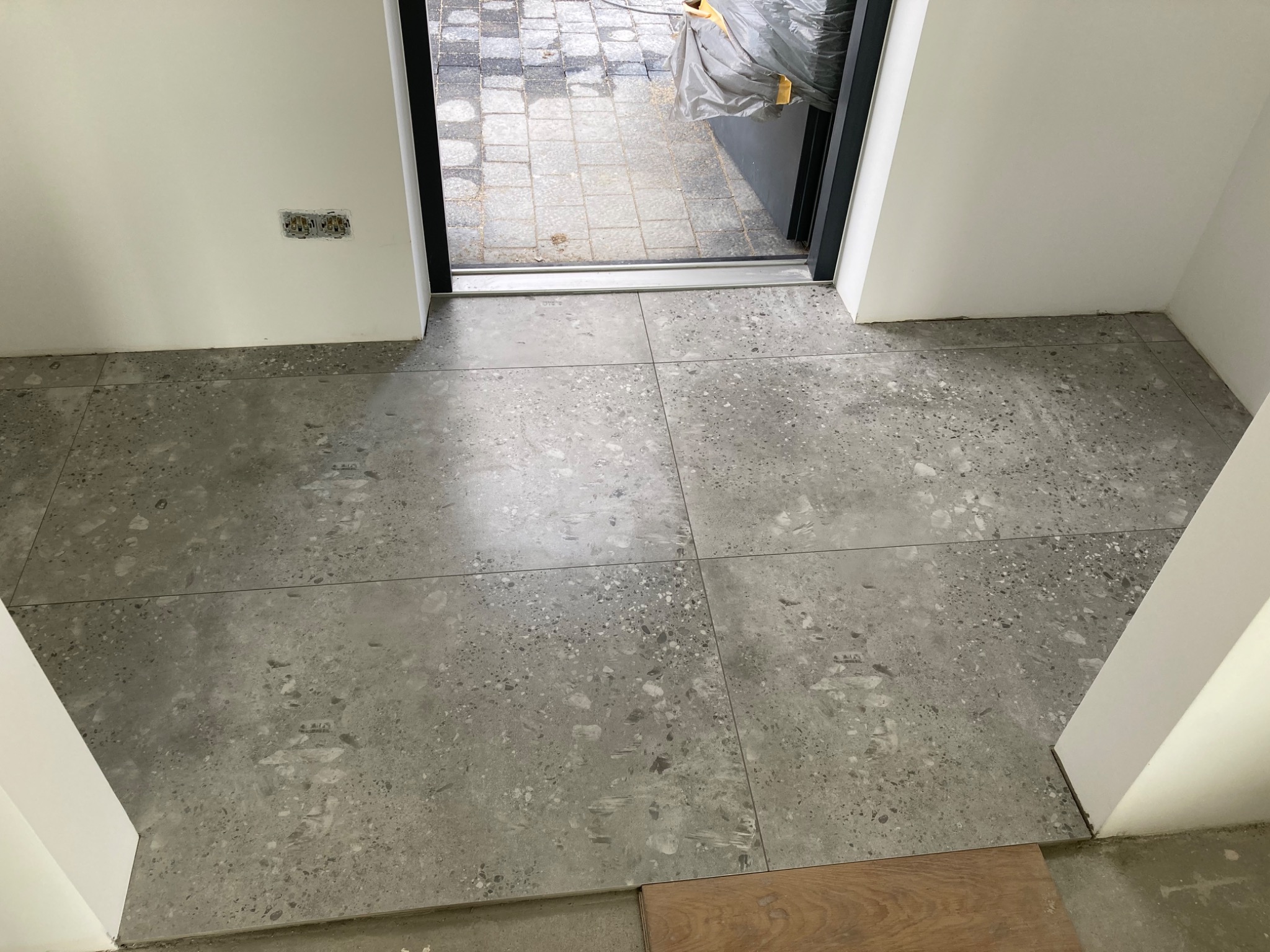 Ułożone szare płytki podłogowe z nakrapianym wzorem, widoczne gniazdka elektryczne bez osłon na ścianie, fragment progu drzwi balkonowych i fragment panela podłogowego.