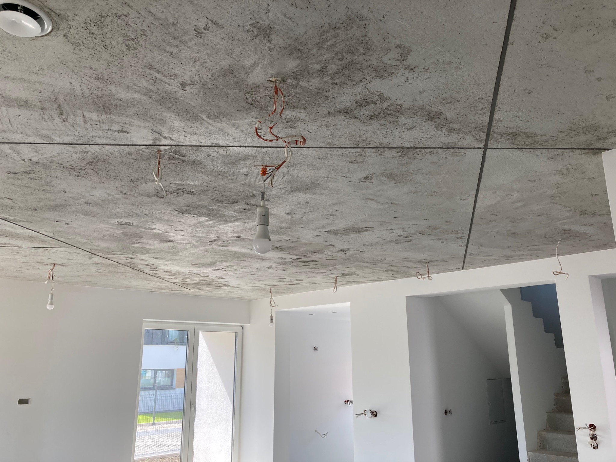 Surowy betonowy sufit w nowym mieszkaniu z widocznymi przewodami elektrycznymi i tymczasową żarówką, białe ściany, okno i widok na schody w głębi.