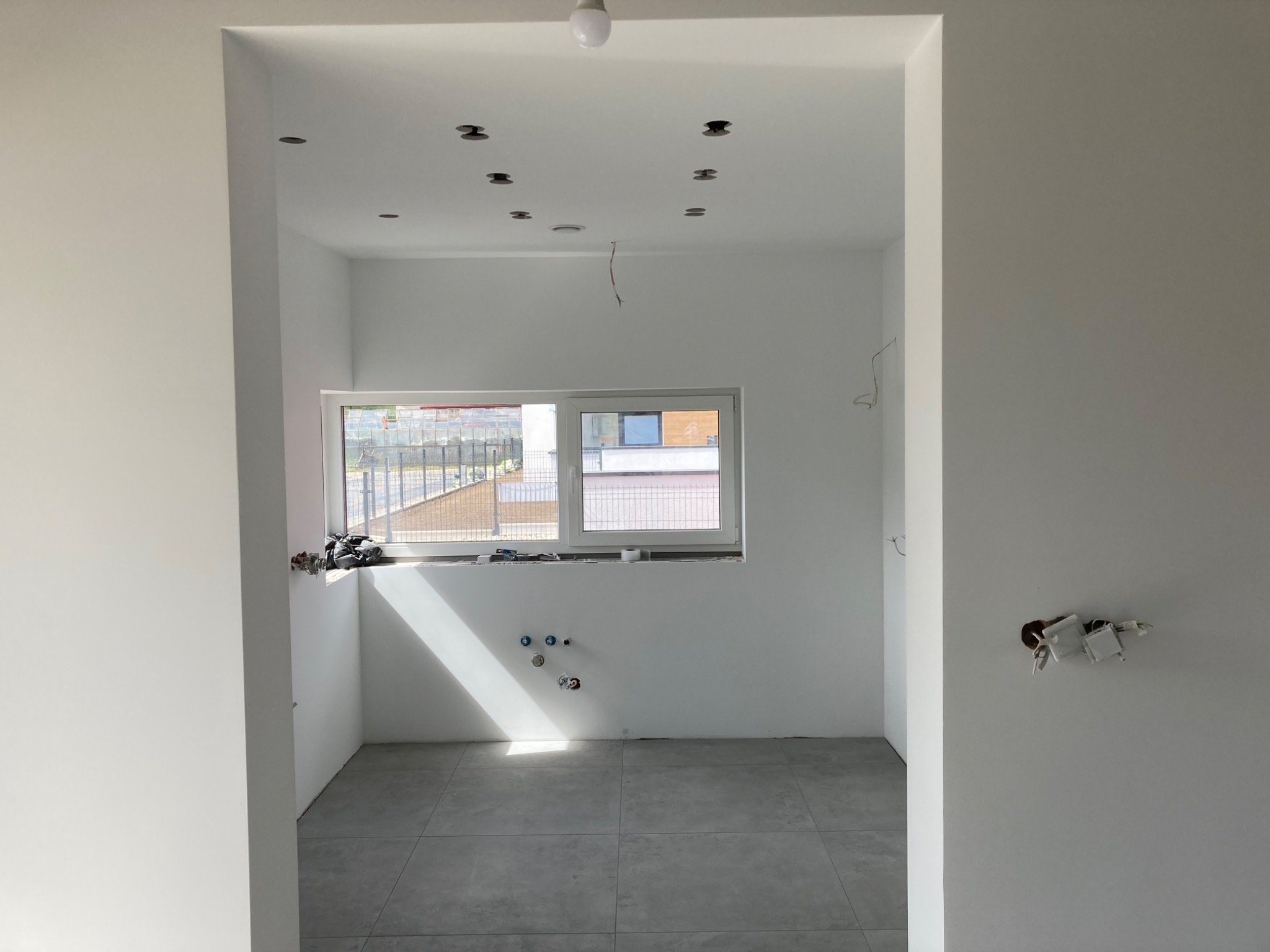 Wnętrze pokoju po remoncie, ściany pomalowane na biało, szara podłoga z płytek, widoczne instalacje elektryczne i hydrauliczne, przygotowane otwory na oświetlenie sufitowe.