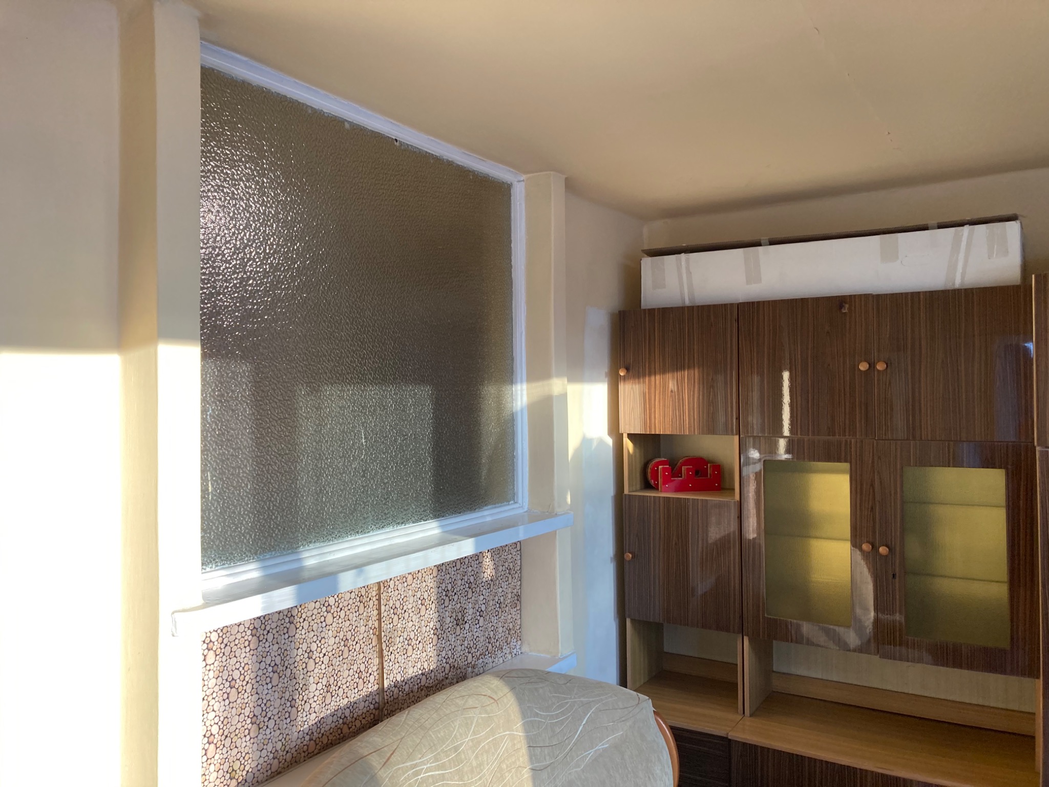 Pokój z oknem z szybą ornamentową, ściany w odcieniach beżu, drewniana boazeria pod oknem i meblościanka z forniru w tle.