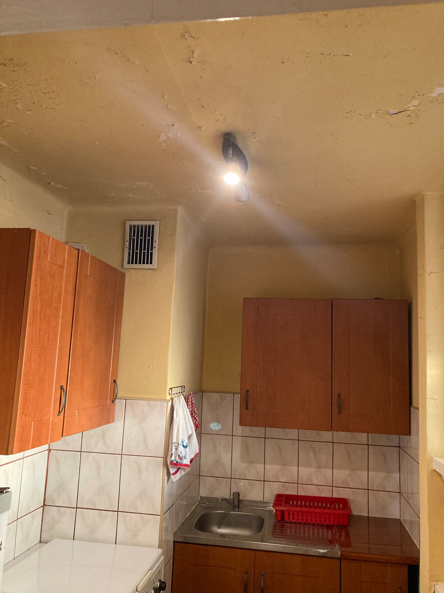 Kuchnia przed remontem: żółty, łuszczący się sufit z lampą, brązowe szafki wiszące nad zlewem i kuchenką, białe płytki na ścianie.