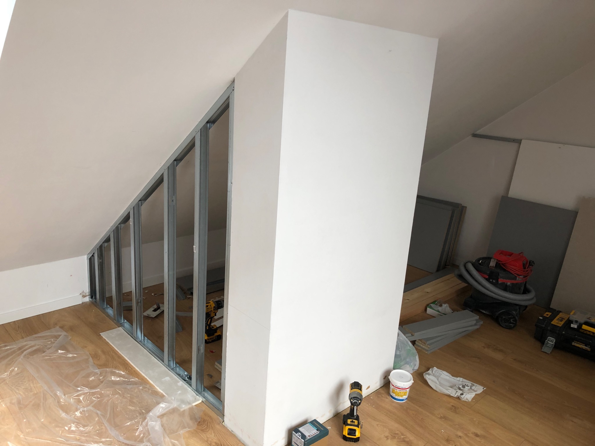 Montaż ścianki działowej z profili stalowych w skosie poddasza, widoczny częściowo zagruntowany fragment ściany, narzędzia pracy i odkurzacz przemysłowy.
