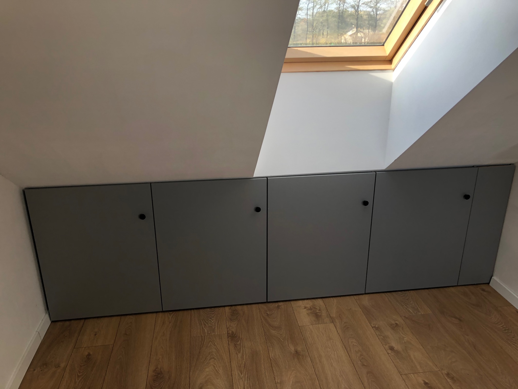 Szare, wbudowane szafki pod skosem dachu, wykończone czarnymi gałkami, na podłodze panele imitujące drewno, widoczne okno dachowe.