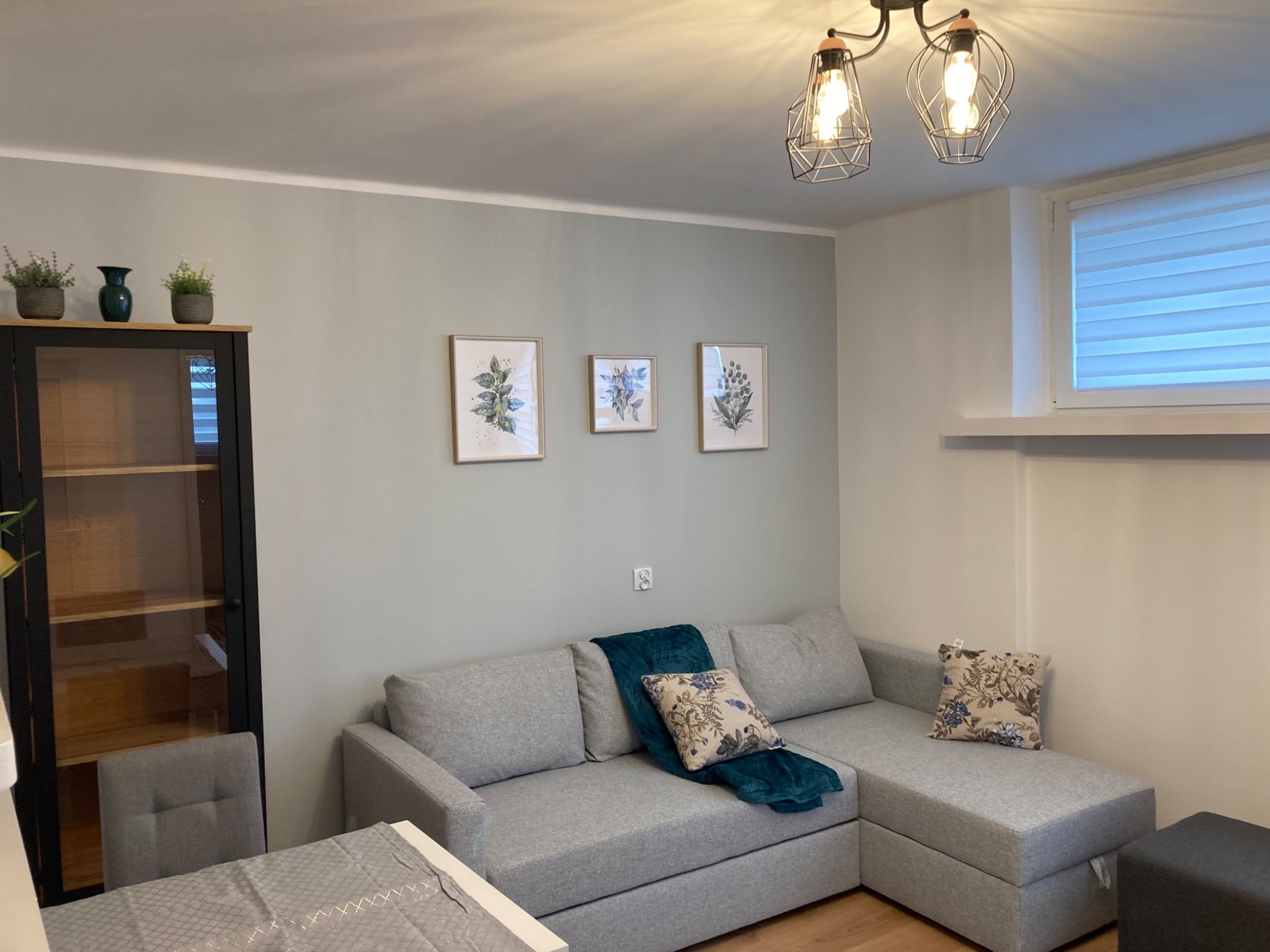 Szary narożnik z turkusowym pledem i poduszkami w kwiaty na tle szaro-zielonej ściany z trzema obrazkami botanicznymi, oświetlony lampą z żarówkami Edisona.