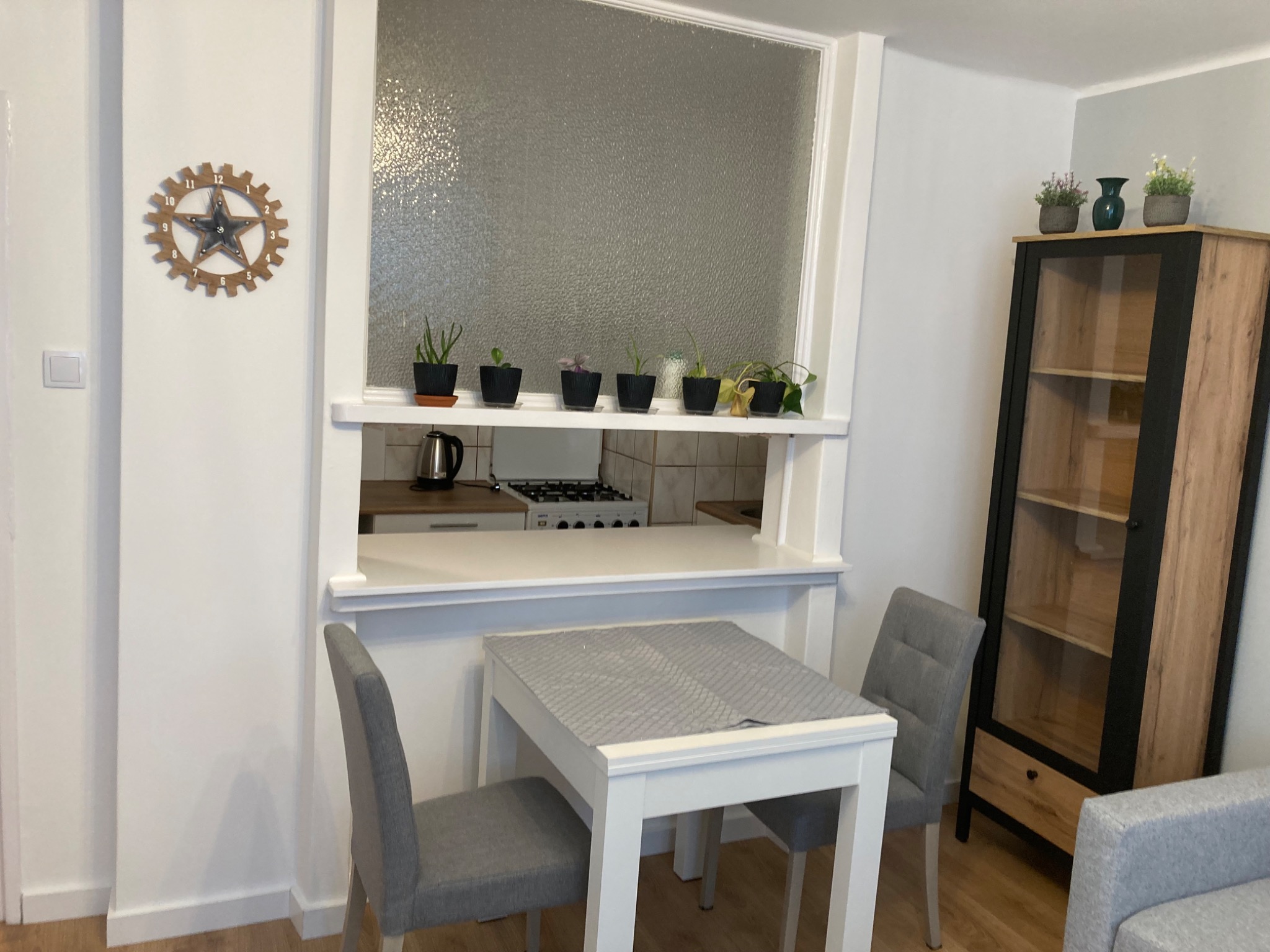 Jasne wnętrze po remoncie: białe ściany, stół z szarymi krzesłami, przeszklona ściana oddzielająca kuchnię z rzędem doniczkowych roślin, dekoracyjny zegar na ścianie.