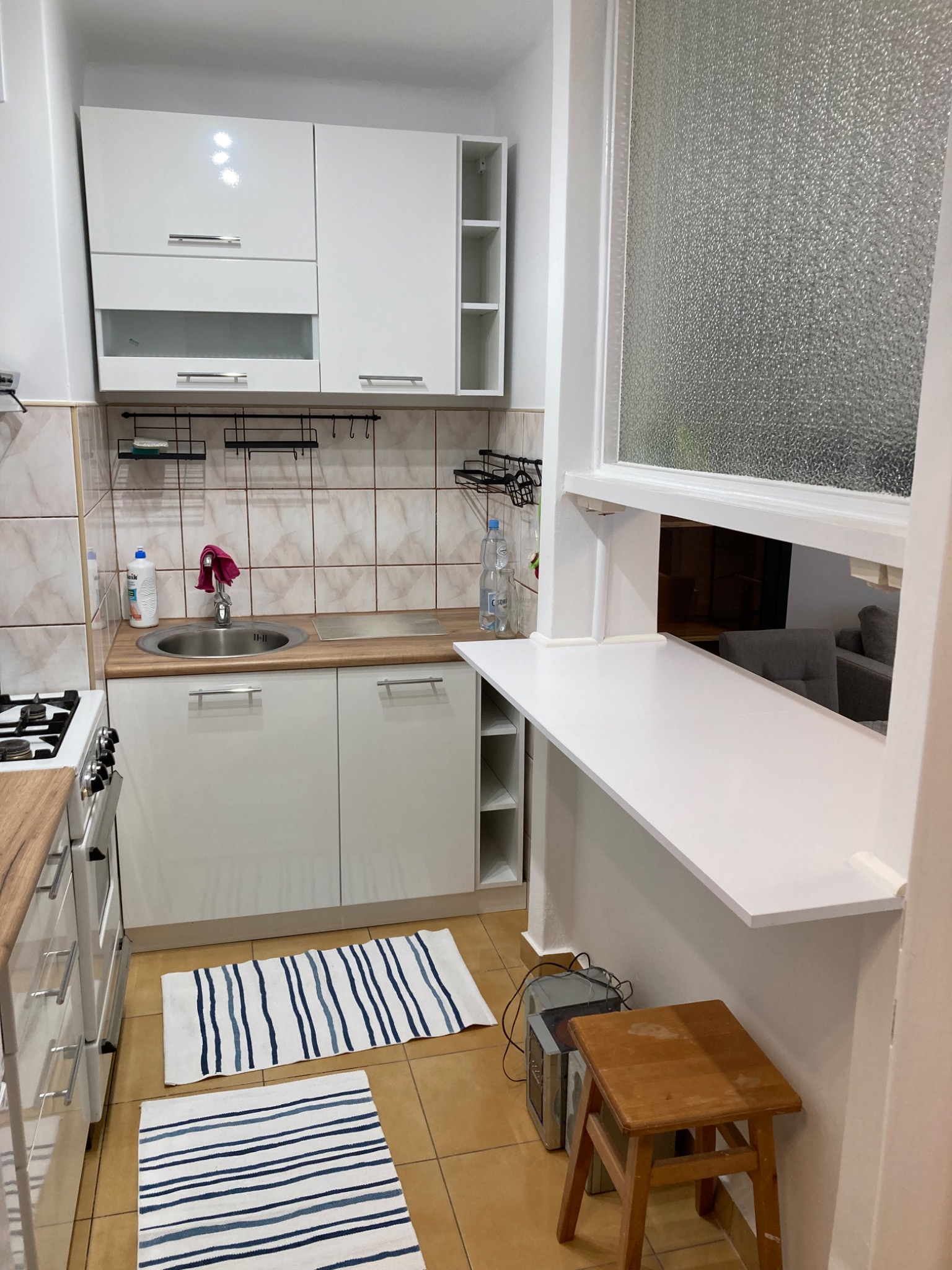 Jasna kuchnia z białymi szafkami, beżowymi płytkami i blatem przechodzącym w okno na salon, widoczne detale jak drewniany stołek i dywaniki w paski.