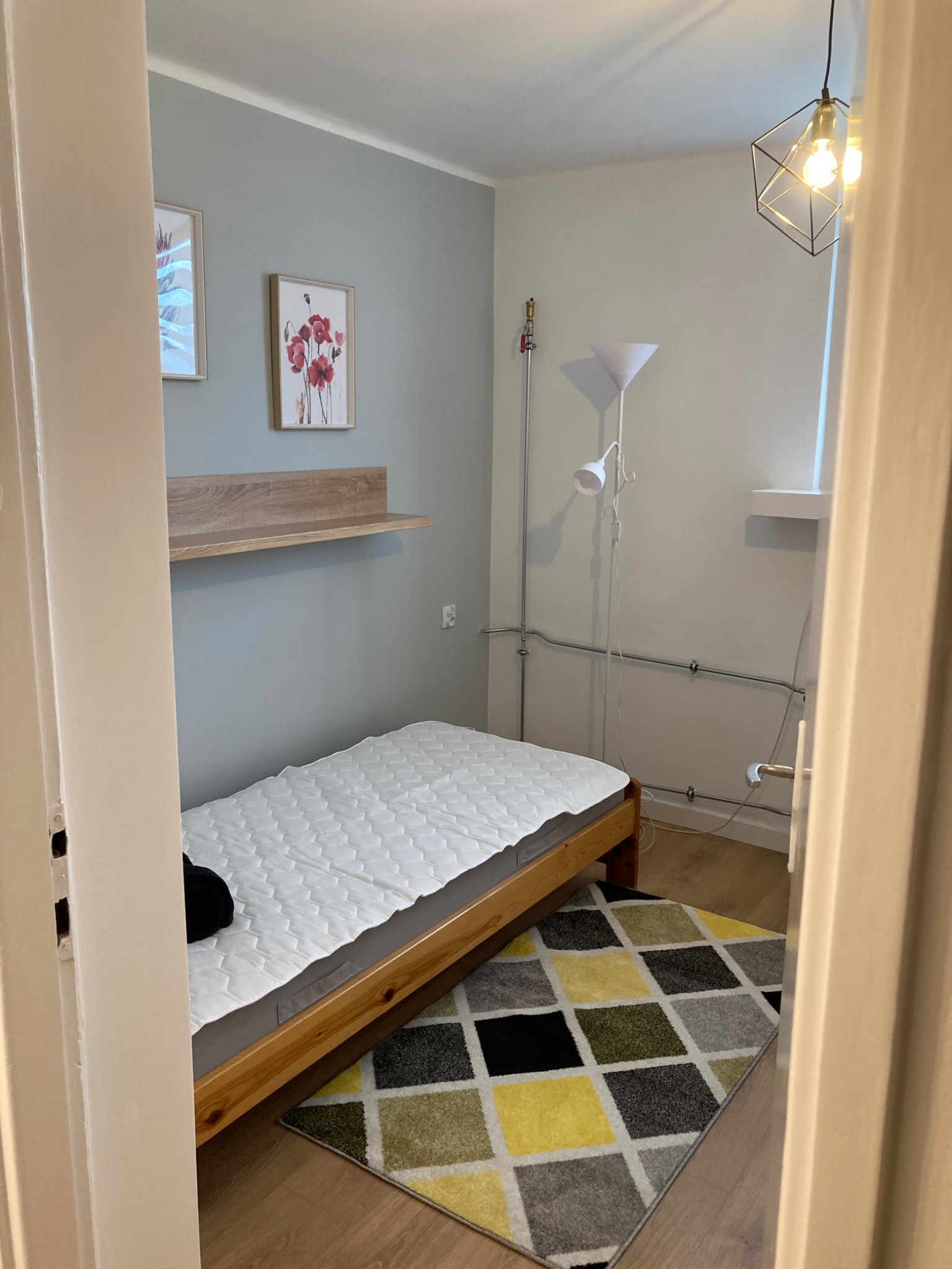 Pokój po remoncie: Ściany pomalowane na szaro, drewniana półka pod obrazami, drewniane łóżko z białym materacem, wzorzysty dywan, lampa wisząca z geometrycznym kloszem, lampa stojąca z białym...