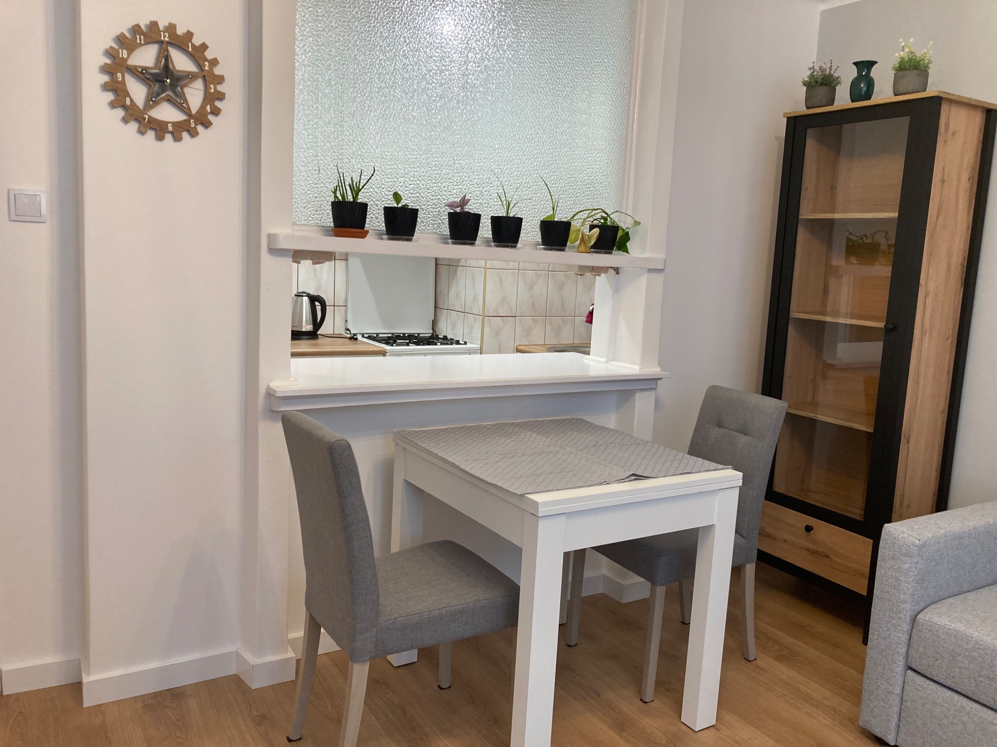 Jasne wnętrze mieszkania z widokiem na kuchnię przez przeszklone okno, stół z krzesłami i dekoracje roślinne.