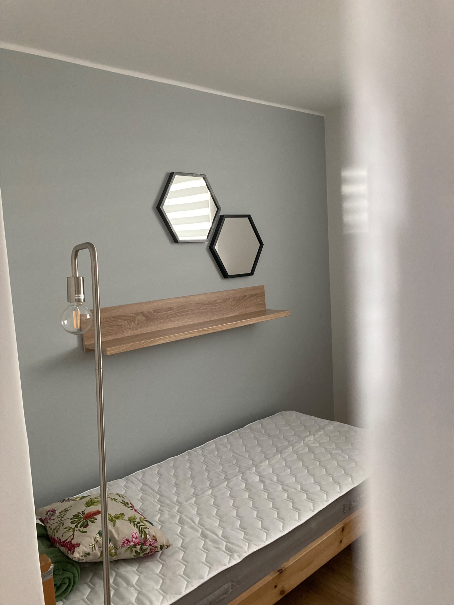 Pokój z szarą ścianą, heksagonalnymi lustrami, drewnianą półką, łóżkiem z materacem i lampą podłogową z żarówką Edisona, widok przez częściowo otwarte drzwi.