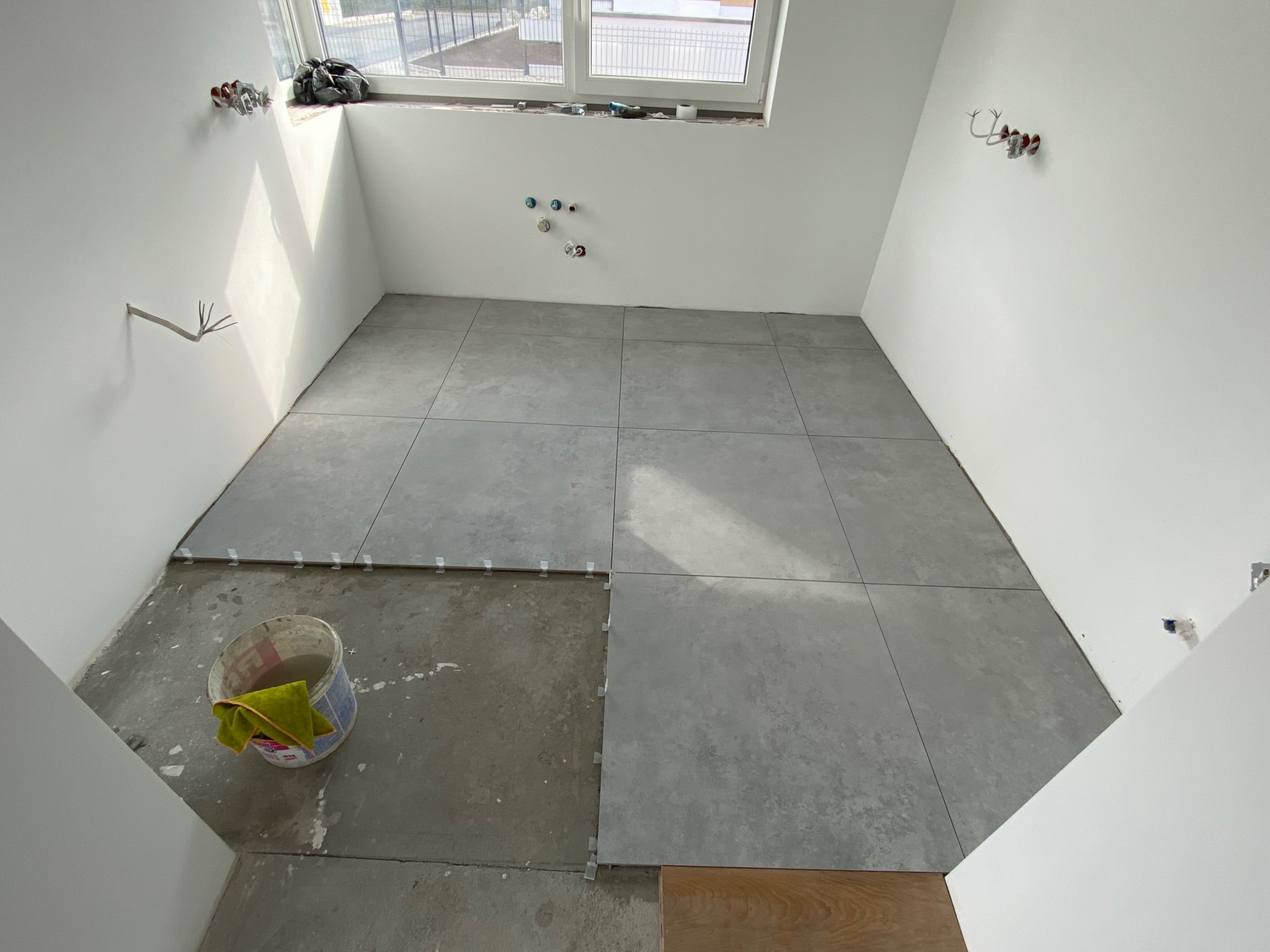 Wnętrze w trakcie układania szarych, kwadratowych płytek podłogowych; widoczny fragment surowej podłogi, plastikowe krzyżyki dystansowe, wiadro z wodą i gąbką oraz wystające kable ze ściany.