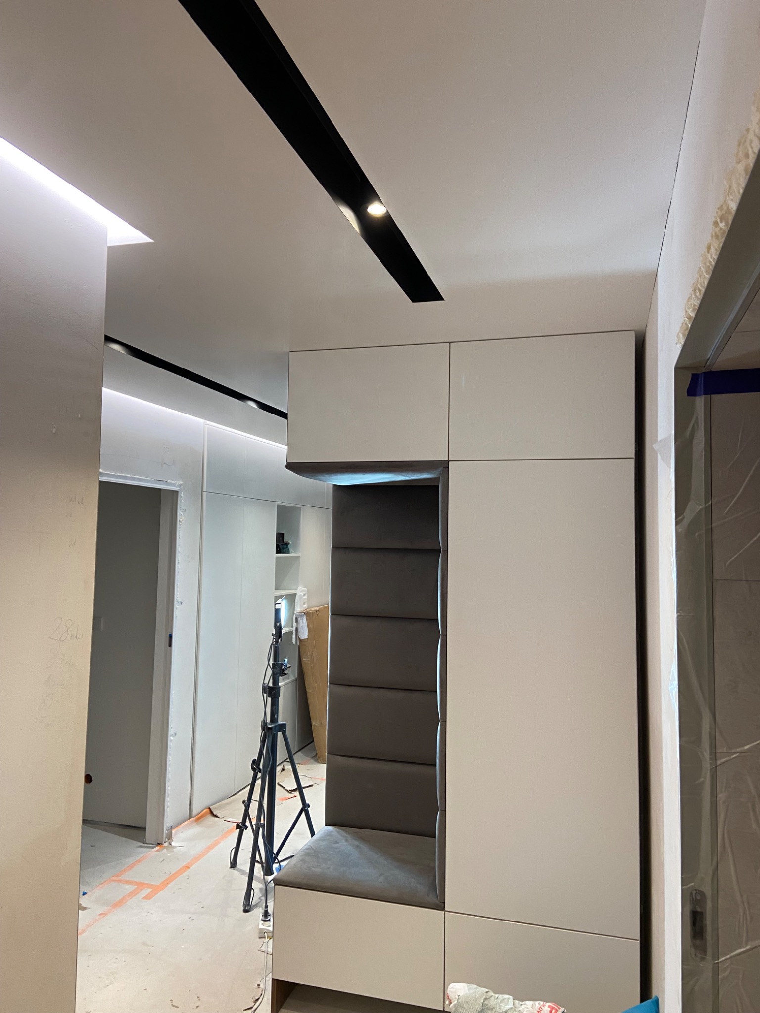 Wnęka z tapicerowanym siedziskiem i oparciem w zabudowie meblowej, widoczny statyw oświetleniowy, sufit z oświetleniem liniowym, ślady prac wykończeniowych na podłodze.