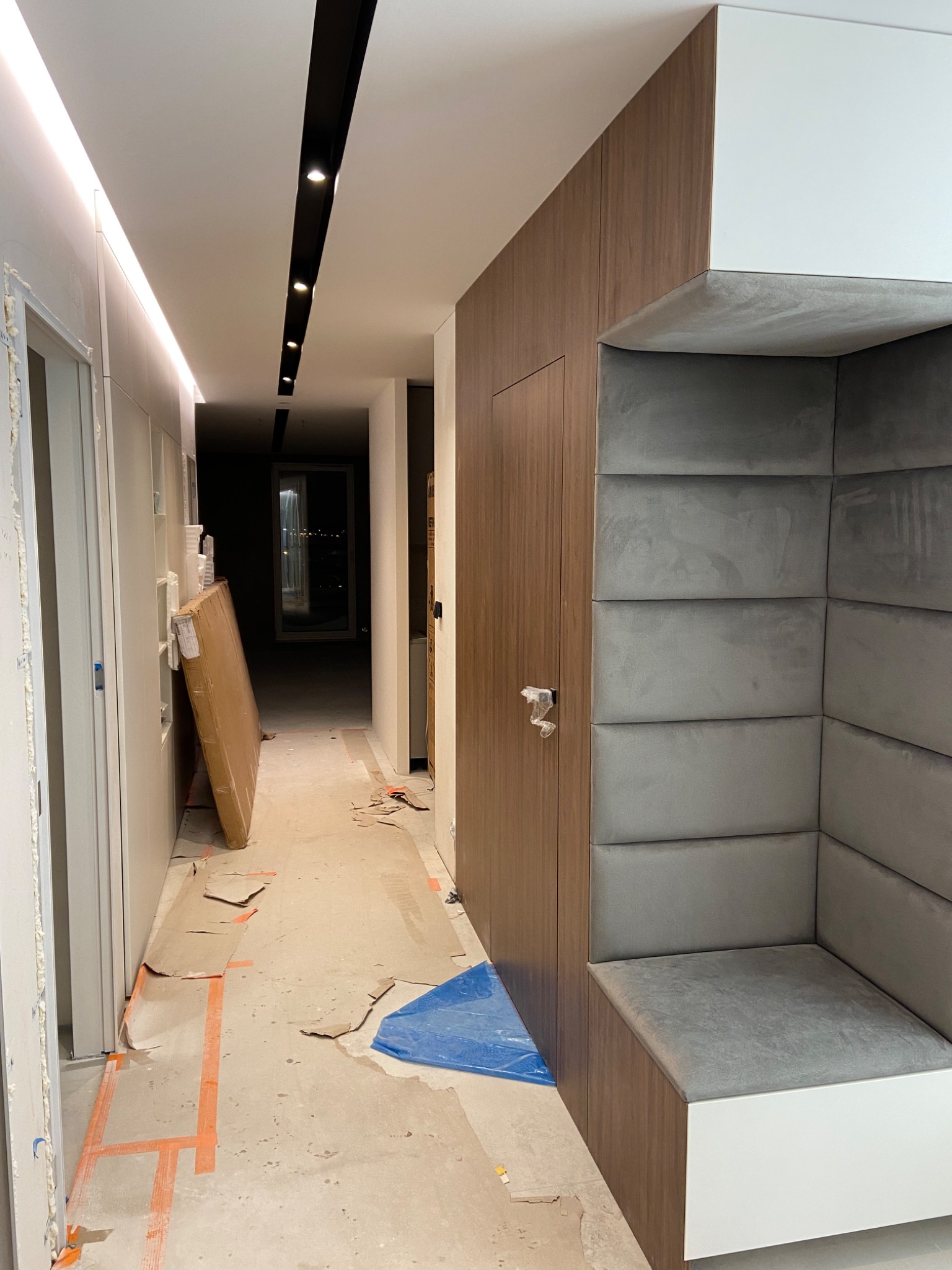 Korytarz w trakcie prac wykończeniowych: zabezpieczona podłoga kartonem i folią, zabudowa z paneli drewnopodobnych z tapicerowanym siedziskiem i ścianą, widoczne oświetlenie sufitowe.