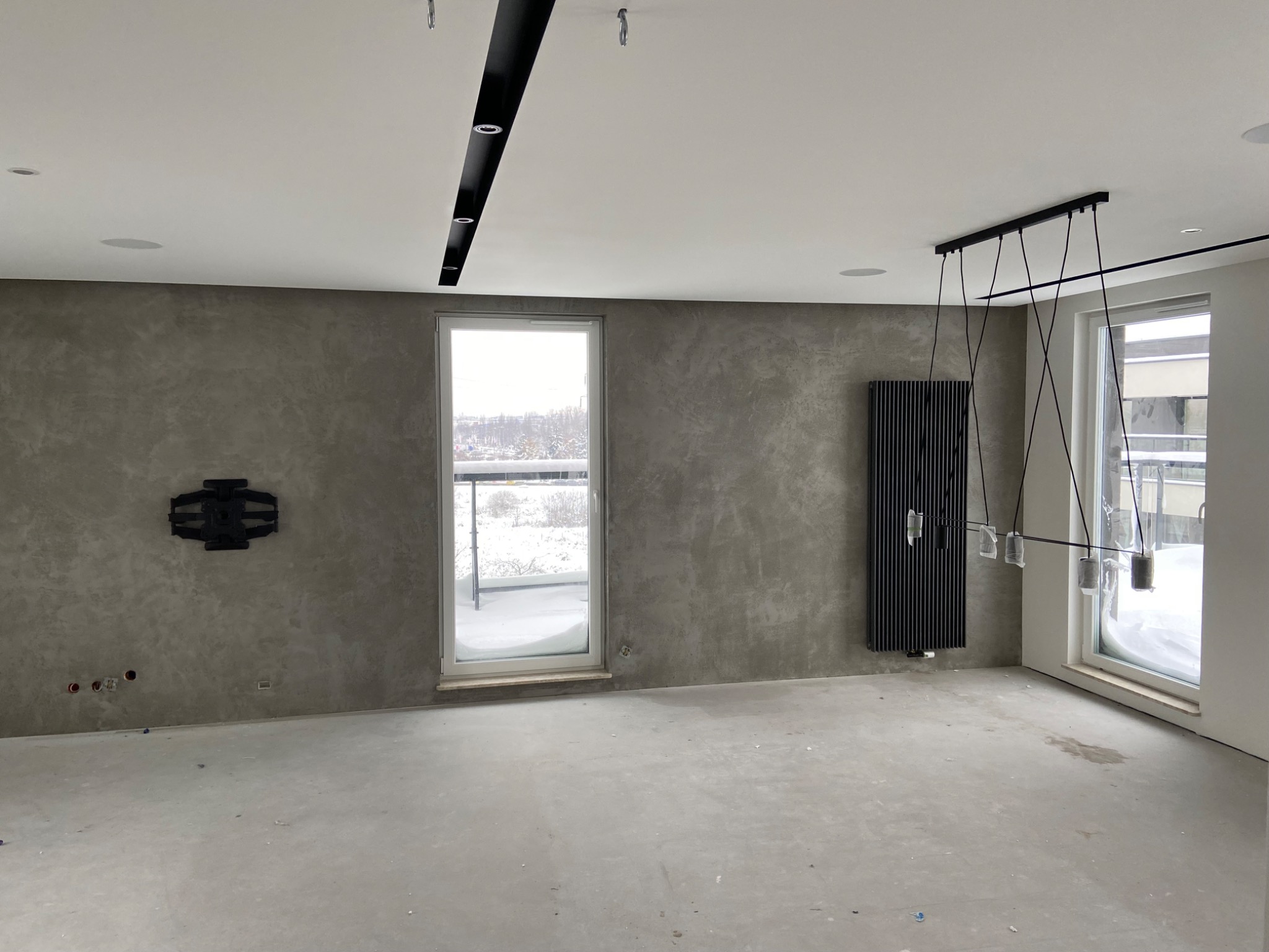 Wnętrze w trakcie wykańczania: ściana pokryta szarą strukturą, nowoczesny czarny grzejnik żeberkowy, sufit z czarną listwą oświetleniową i wisząca lampa z transparentnymi kloszami, gniazdka...