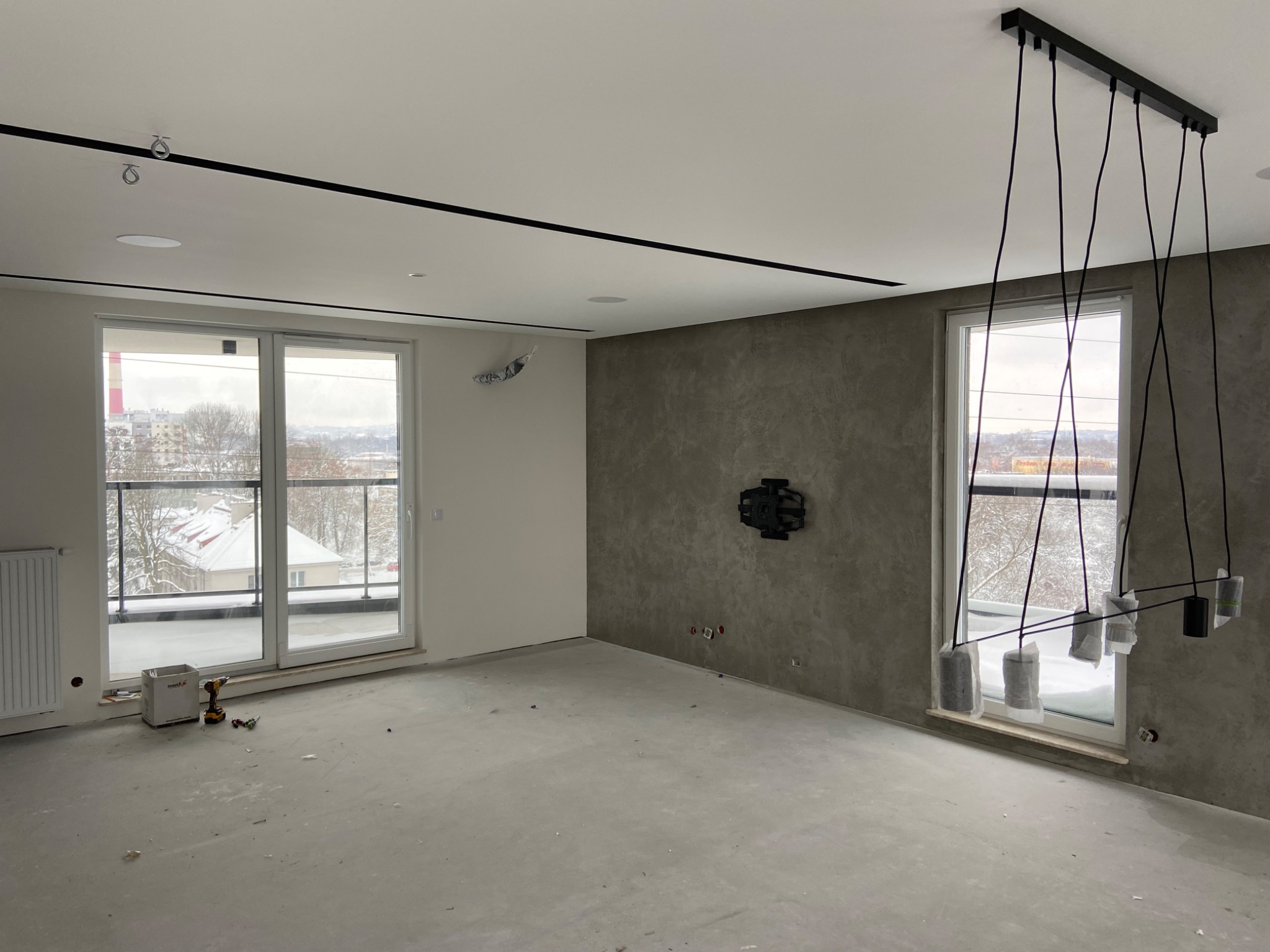 Wnętrze w trakcie remontu: surowa podłoga, ściana z efektem betonu, okna z widokiem na zaśnieżone miasto, nowoczesna lampa wisząca z osłonami, narzędzia do montażu przy oknie balkonowym.
