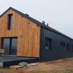 ''P.P.U.H. Kmp Jacek Pazdan'' - Nowoczesny dom jednorodzinny z fasadą z pionowych desek w odcieniach drewna i grafitową elewacją z oknami, widok z zewnątrz na taras z krzesłami i stolikiem nakrytym kwiecistym obrusem, w tle drugi...