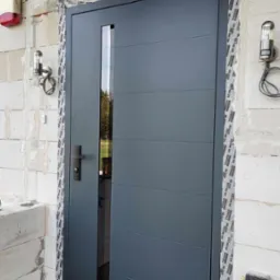 Szare drzwi zewnętrzne z wąskim przeszkleniem, zamontowane w surowym murze z gazobetonu, zabezpieczone taśmą uszczelniającą, widoczne lampy zewnętrzne i fragment instalacji.