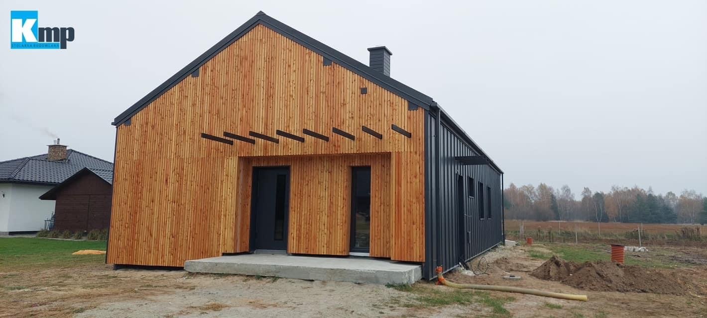 Nowoczesna stodoła z drewnianą elewacją i czarnymi akcentami, okna i drzwi w kolorze antracytowym, betonowy podest wejściowy, widoczne ślady prac ziemnych wokół budynku.