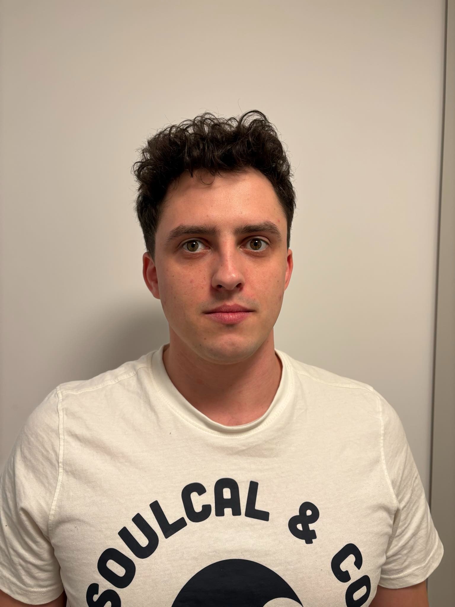 Portret młodego mężczyzny z krótkimi, kręconymi włosami, ubranego w białą koszulkę z nadrukiem na tle jasnej ściany.