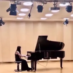 Kobieta w czarnej sukni gra na czarnym fortepianie w sali z oświetleniem scenicznym.