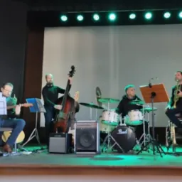 Zespół muzyczny na scenie podczas występu: gitarzysta z nutami, kontrabasista, perkusista Pearl i trębacz siedzący na krześle obok statywu z nutami.