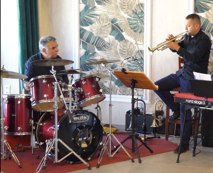 Dwaj muzycy podczas występu: perkusista gra na czerwonej perkusji marki Pacific, a trębacz gra na trąbce, siedząc na krześle obok keyboardu Nord Stage 3.