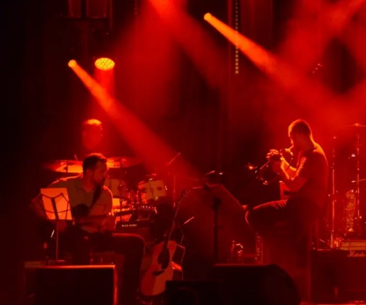 Zespół instrumentalny podczas występu na scenie oświetlonej czerwonymi reflektorami: gitarzysta z nutami, trębacz i perkusista w tle.