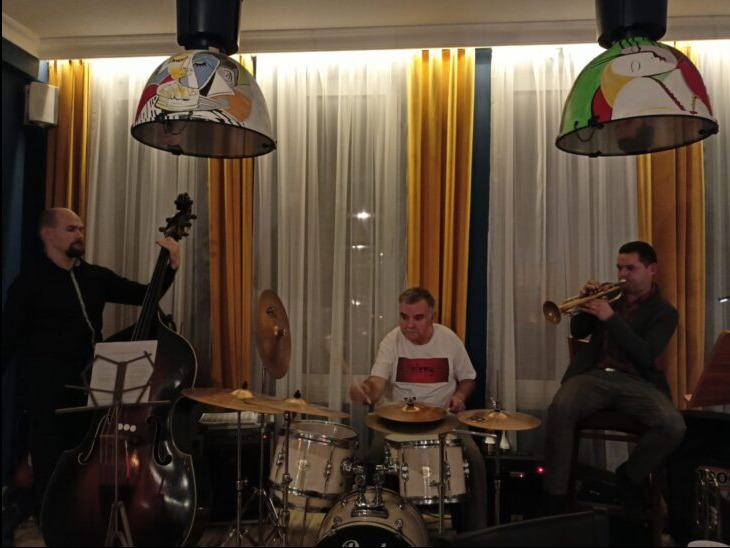 Jazzowy tercet w trakcie występu: kontrabasista, perkusista i trębacz grają w pomieszczeniu z designerskimi lampami wiszącymi z motywami Picassa.