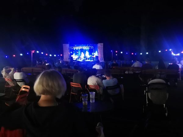 Widok z tyłu na publiczność siedzącą na krzesłach i ławkach podczas koncertu na zewnątrz, scena oświetlona niebieskimi światłami, girlandy z kolorowymi żarówkami nad głowami widzów, w tle ciemne...