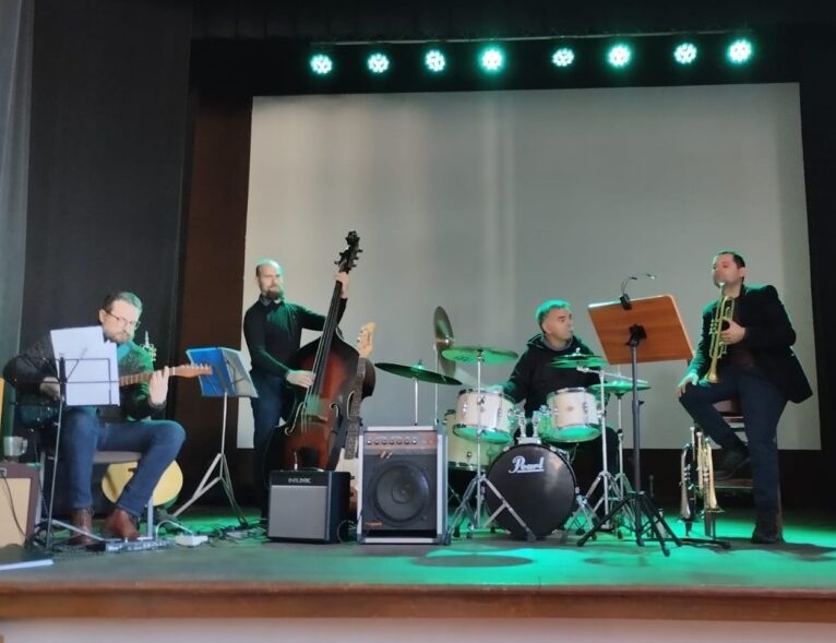 Zespół muzyczny na scenie podczas występu: gitarzysta z nutami, kontrabasista, perkusista Pearl i trębacz siedzący na krześle obok statywu z nutami.