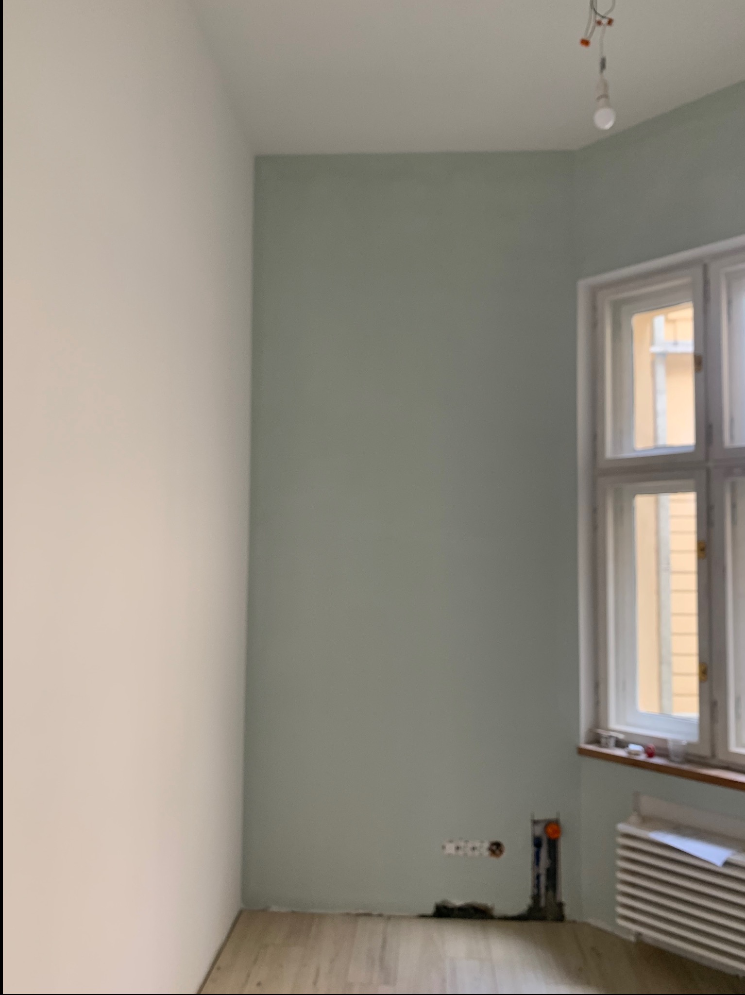 Pokój po częściowym odświeżeniu: jedna ściana biała, druga pomalowana na odcień zieleni, widoczne gniazdka elektryczne i rury przy podłodze, okno z białymi ramami i grzejnik pod nim.