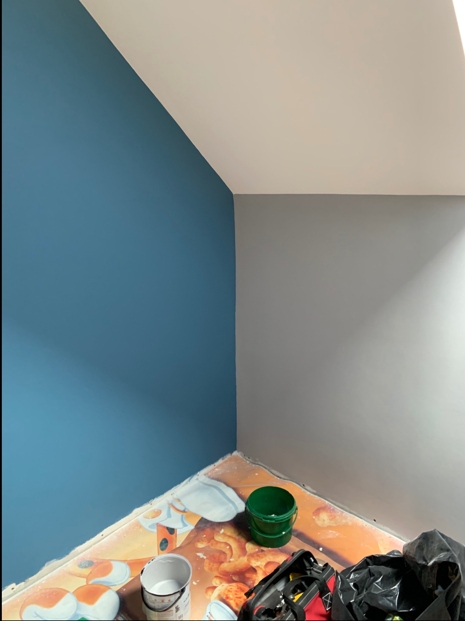 Pomieszczenie po częściowym pomalowaniu ścian na kolor niebieski i szary, z widocznymi narzędziami malarskimi i resztkami farby na podłodze z wzorem pieczywa.