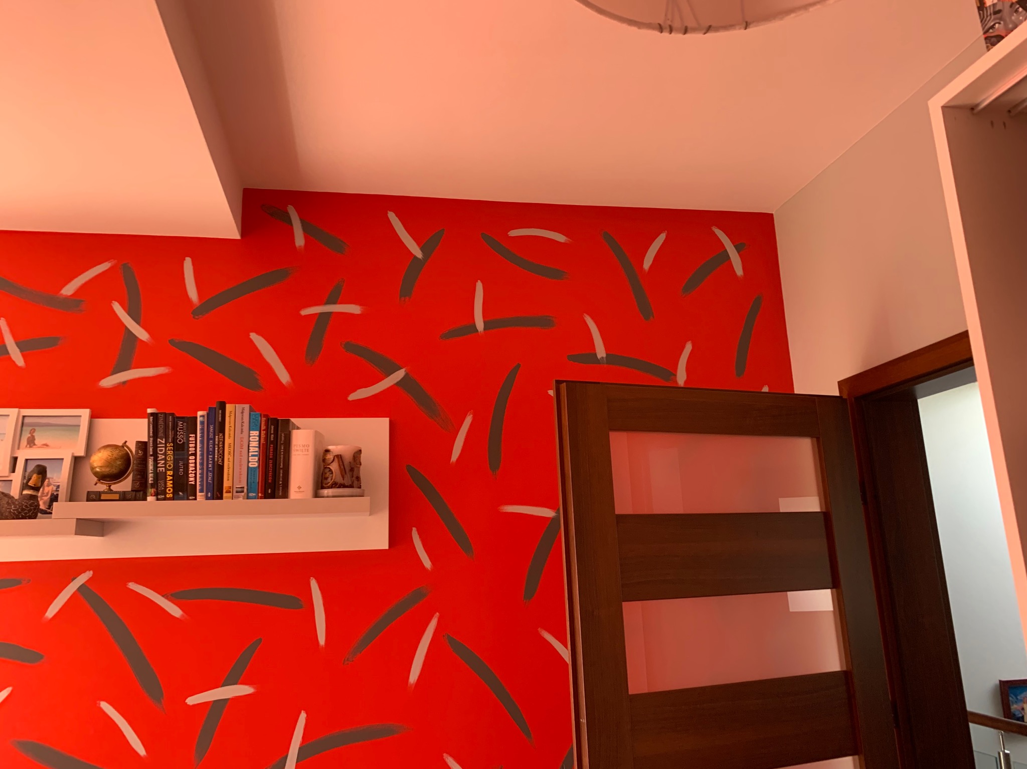 Czerwona ściana z abstrakcyjnym wzorem w odcieniach szarości i bieli, półka z książkami, globusem i ramkami ze zdjęciami, brązowe drzwi z przeszkleniami.